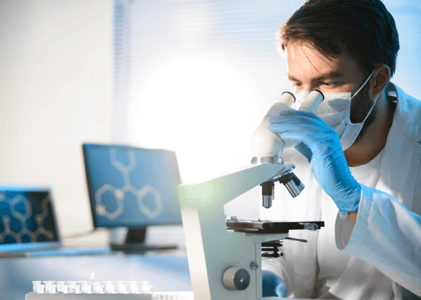 Male scientist wearing a mask and gloves looking into a microscope with molecular diagrams on monitors behind him