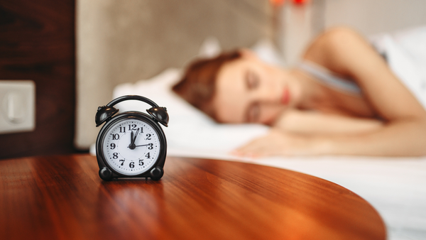 Alarm clock on a bedside table with a woman sleeping in bed in the background