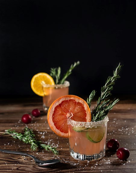 Two festive mocktails garnished with citrus slices and rosemary on a wooden table with cranberries