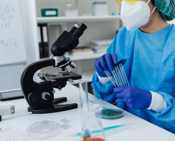 Lab scientist in protective gown and mask holding test tubes beside a microscope