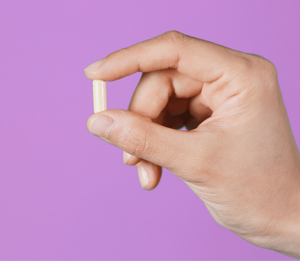 Hand holding a single beige capsule against a purple background