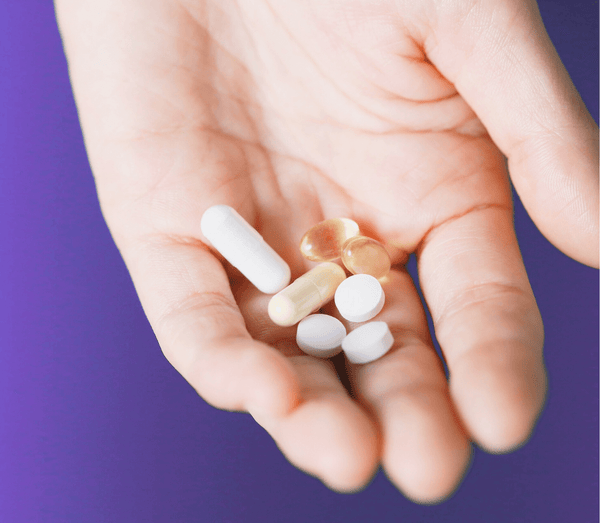 Hand holding an assortment of white and gold supplement capsules against a purple background