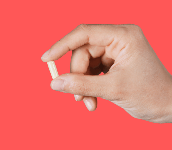 Hand holding a single beige capsule against a coral background