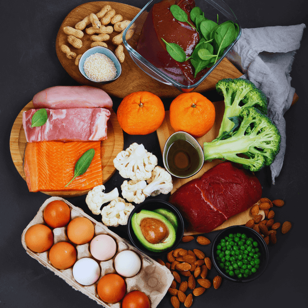 Overhead spread of protein-rich foods including liver, salmon, ham, eggs, broccoli and almonds