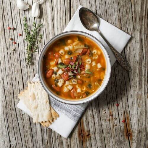 Overhead view of a bowl of chunky minestrone soup with pasta and vegetables, served with crackers and a spoon