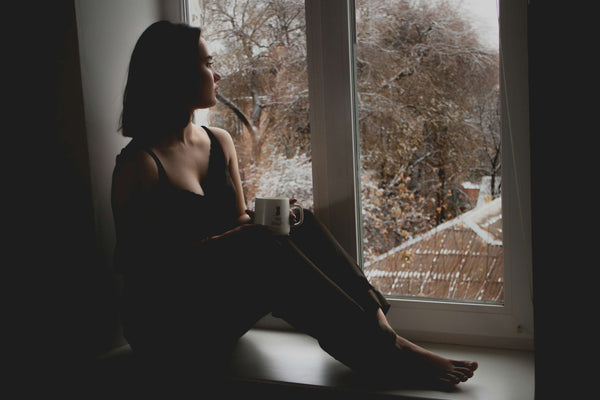 Woman sitting on a windowsill holding a mug and looking out at a snowy scene