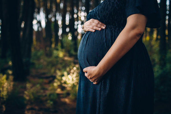 Close-up of a pregnant woman's belly in a navy striped dress, hands cradling the bump in a forest setting