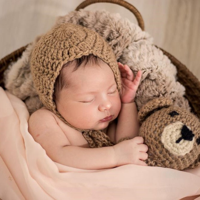 Sleeping newborn in a knitted bonnet cuddling a crochet teddy bear in a basket