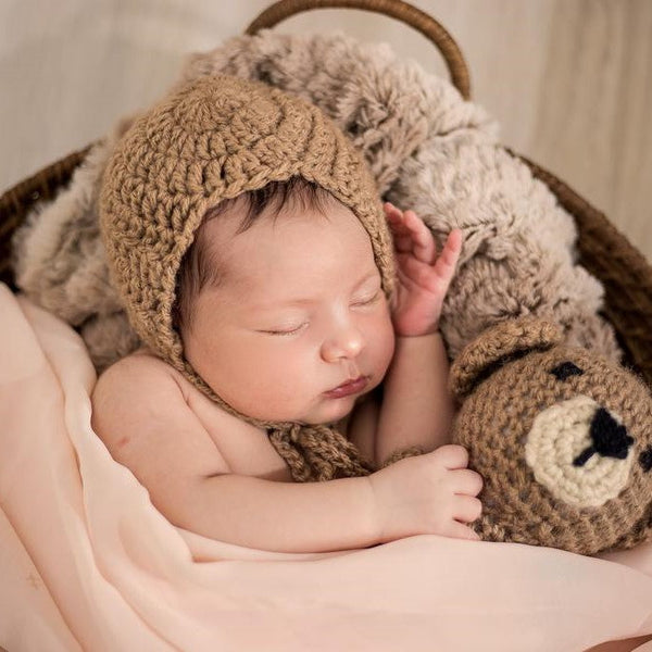 Sleeping newborn in a knitted bonnet cuddling a crochet teddy bear in a basket