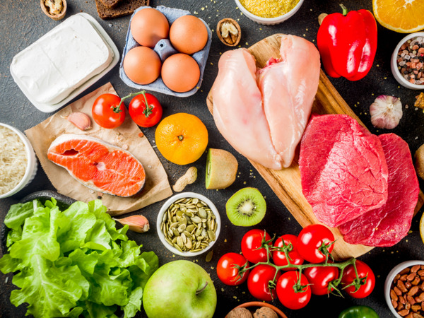 Overhead view of raw meat, poultry, fish, eggs, vegetables and nuts arranged on a dark surface