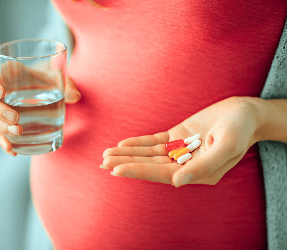 Close-up of a pregnant woman's belly in red holding a handful of supplement capsules and a glass of water