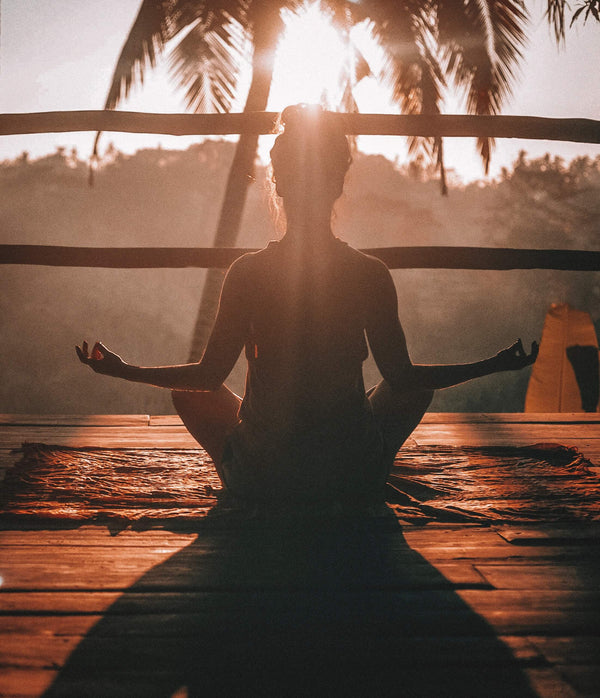Silhouette of a woman meditating cross-legged on a deck at sunrise beneath palm trees