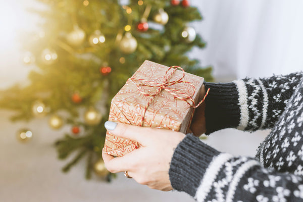 Hands in a knitted jumper holding a wrapped Christmas gift in front of a decorated tree