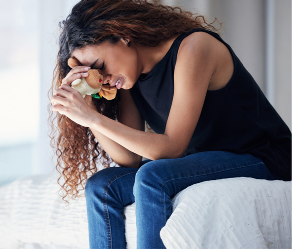 Woman sitting on a bed holding a small stuffed toy to her face while crying