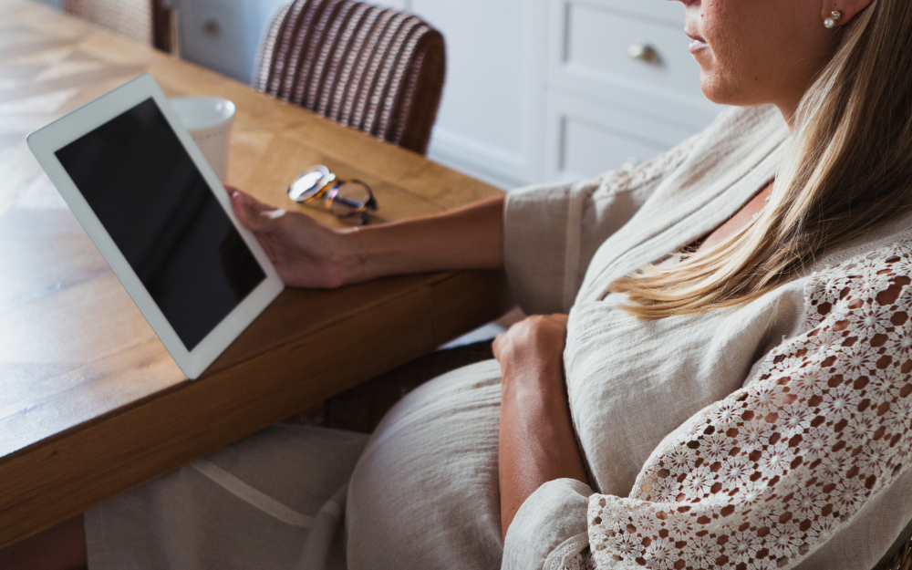 Pregnant woman sitting at a table with a tablet device and a cup of coffee