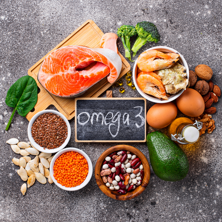 Omega-3 rich foods including salmon, shrimp, flaxseeds, lentils, eggs, avocado, walnuts and broccoli arranged around a chalkboard sign reading 'omega 3'