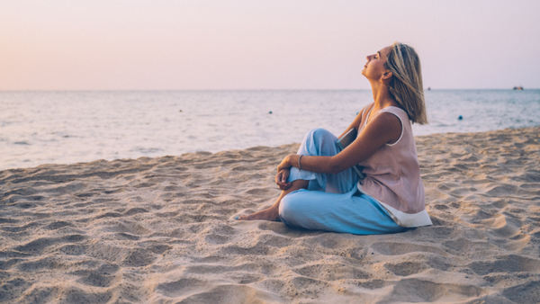Woman sitting cross-legged on a beach at sunset looking up at the sky