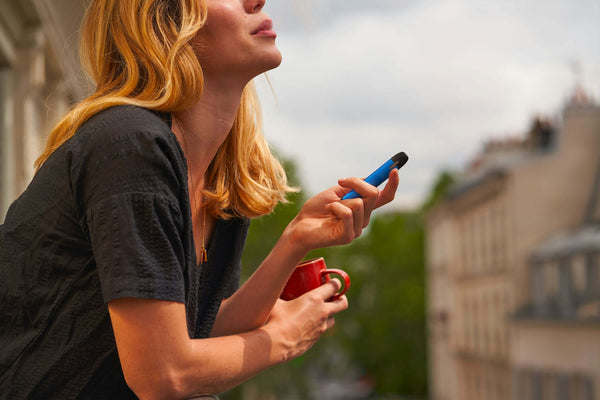Woman on a balcony holding a vape pen and a red mug overlooking a city
