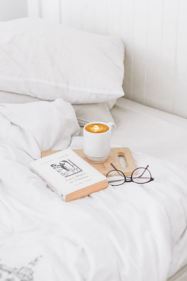 Cup of coffee, a book and glasses resting on a tray on a white bed
