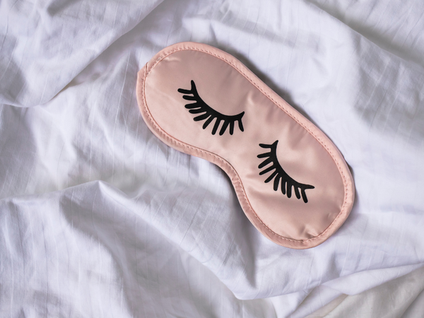 Pink sleep mask with embroidered closed eyelashes resting on a bed