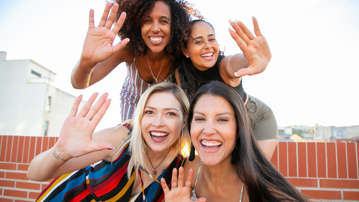 Four smiling women waving at the camera on a rooftop on a sunny day