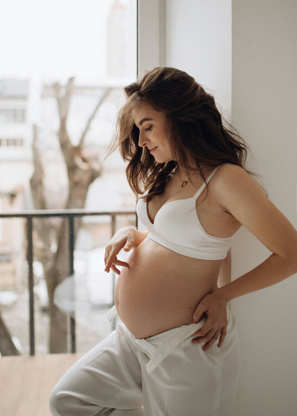 Pregnant woman in white leaning against a window smiling down at her belly