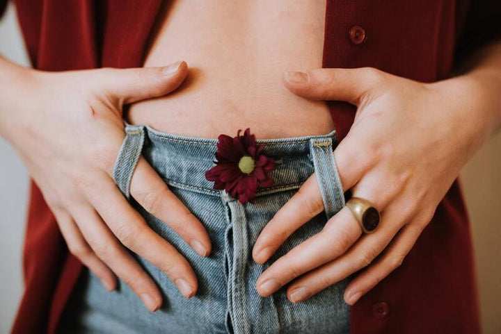 Person holding their lower abdomen, with a dark red daisy tucked into the waistband of light blue jeans