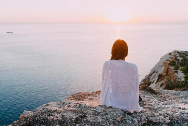 Woman sitting on coastal cliff rocks, gazing out at a calm sea during sunset