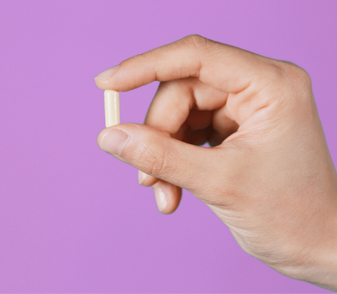 Hand holding a single beige capsule against a purple background