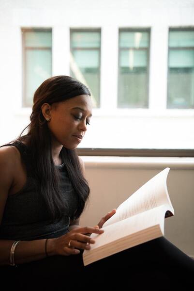 Woman sitting by a window reading a book intently, with long dark hair and a sleeveless top