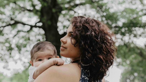 Woman holding her baby over her shoulder outdoors beneath trees