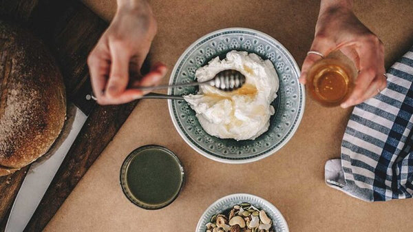 Overhead shot of hands drizzling honey onto a bowl of yogurt beside sourdough bread and a green juice