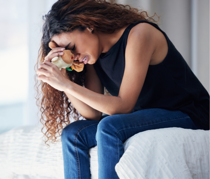 Woman sitting on a bed, hunched over and crying while holding a small soft toy