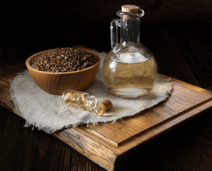 Flaxseeds in a wooden bowl beside a glass bottle of oil and small gel capsules on a linen cloth