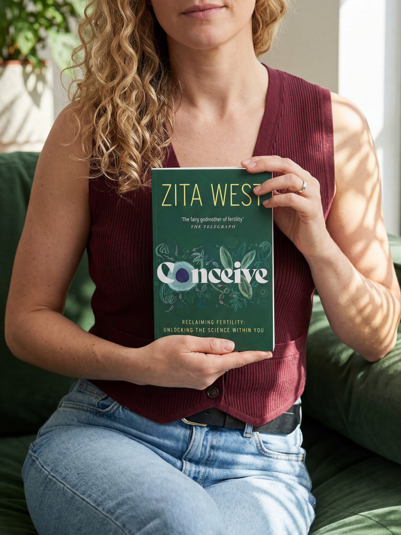 Person holding a book titled 'Conceive' by Zita West on a green couch.