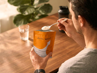 A man in a gray t-shirt is shown from behind, holding a Zita West Vitaseed Pro pouch and using a spoon to scoop white powder. He is seated at a wooden table with a glass of water and a shaker bottle in the background.