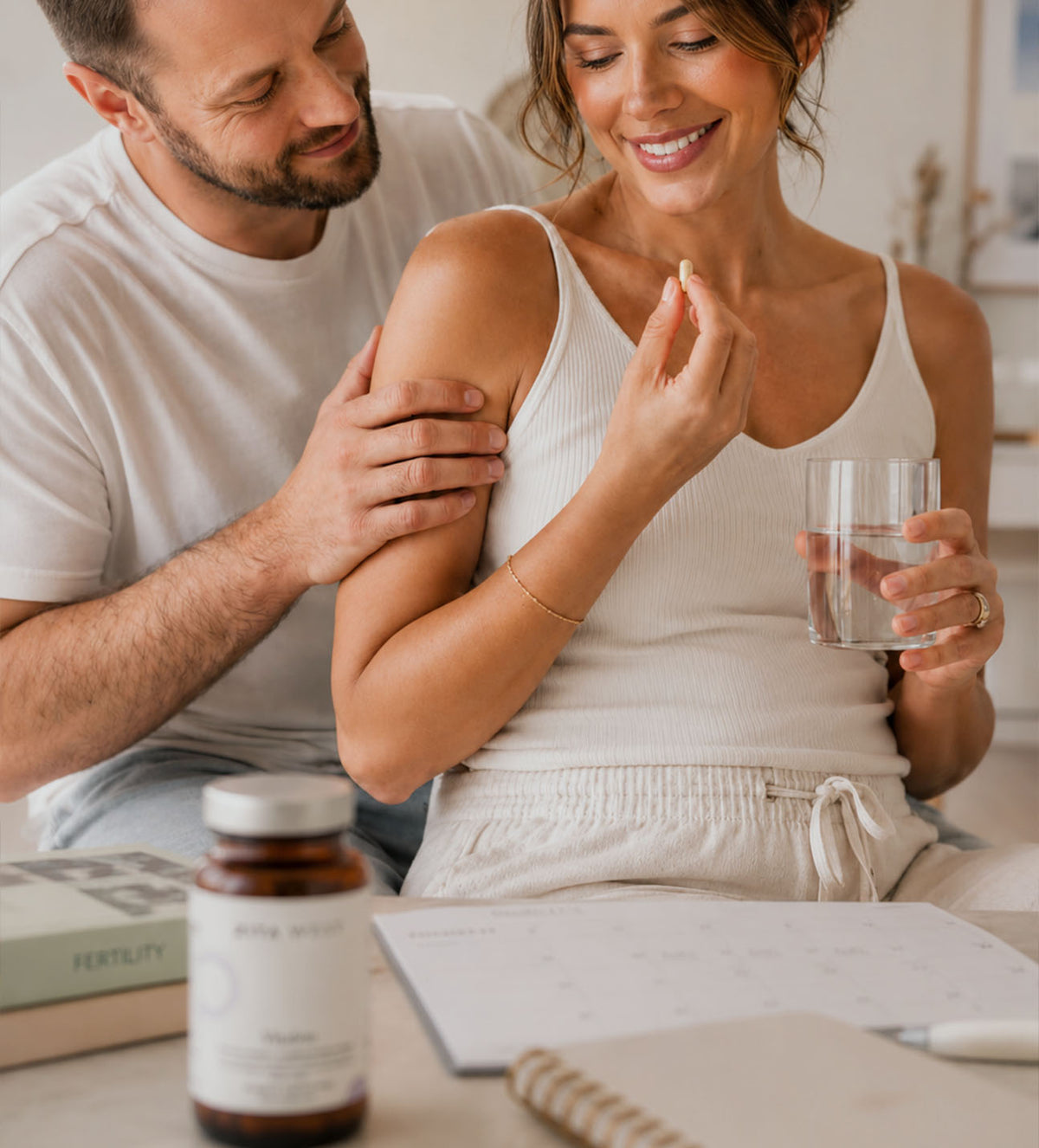 A smiling woman with a man behind her. She is taking her daily supplement